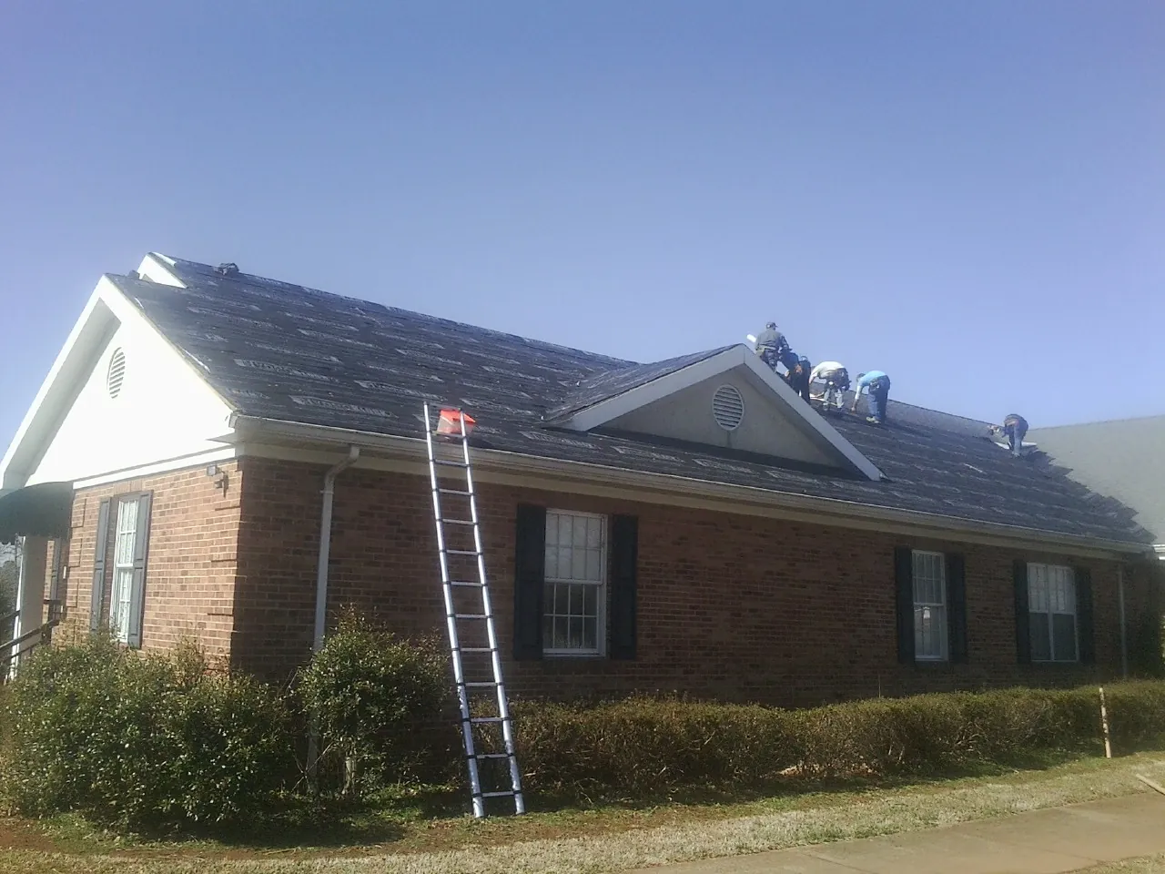 Crew tearing shingles off a brick ranch roof with a ladder set — Metal Roof Repair work by our crew in Loranger
