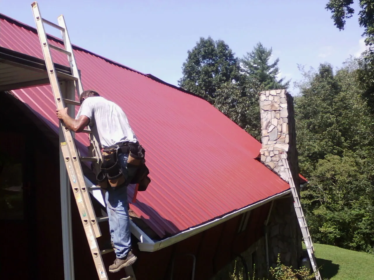 Roofer on a ladder beside a red metal roof and stone chimney — Soffit & Fascia Repair by Roofing Brothers in Loranger